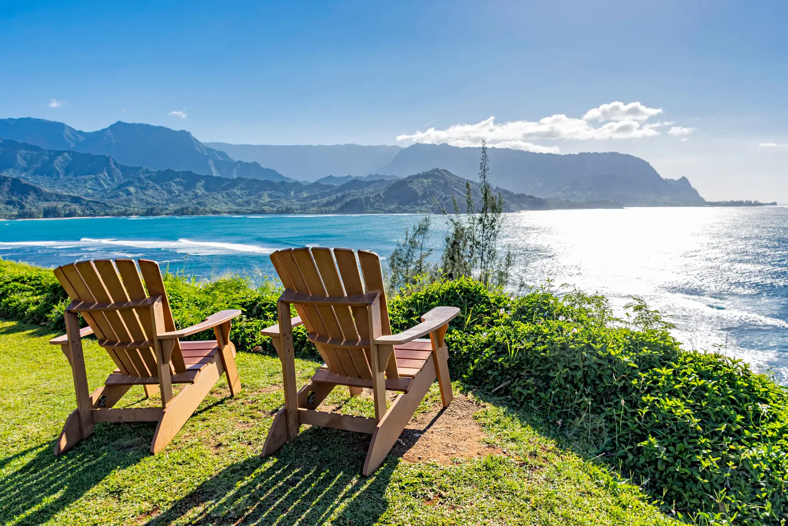 Ocean and mountain views on Kauaʻi's North Shore. Two wooden Adirondack chairs on a green lawn face the ocean, with a palm and mountains across the water.