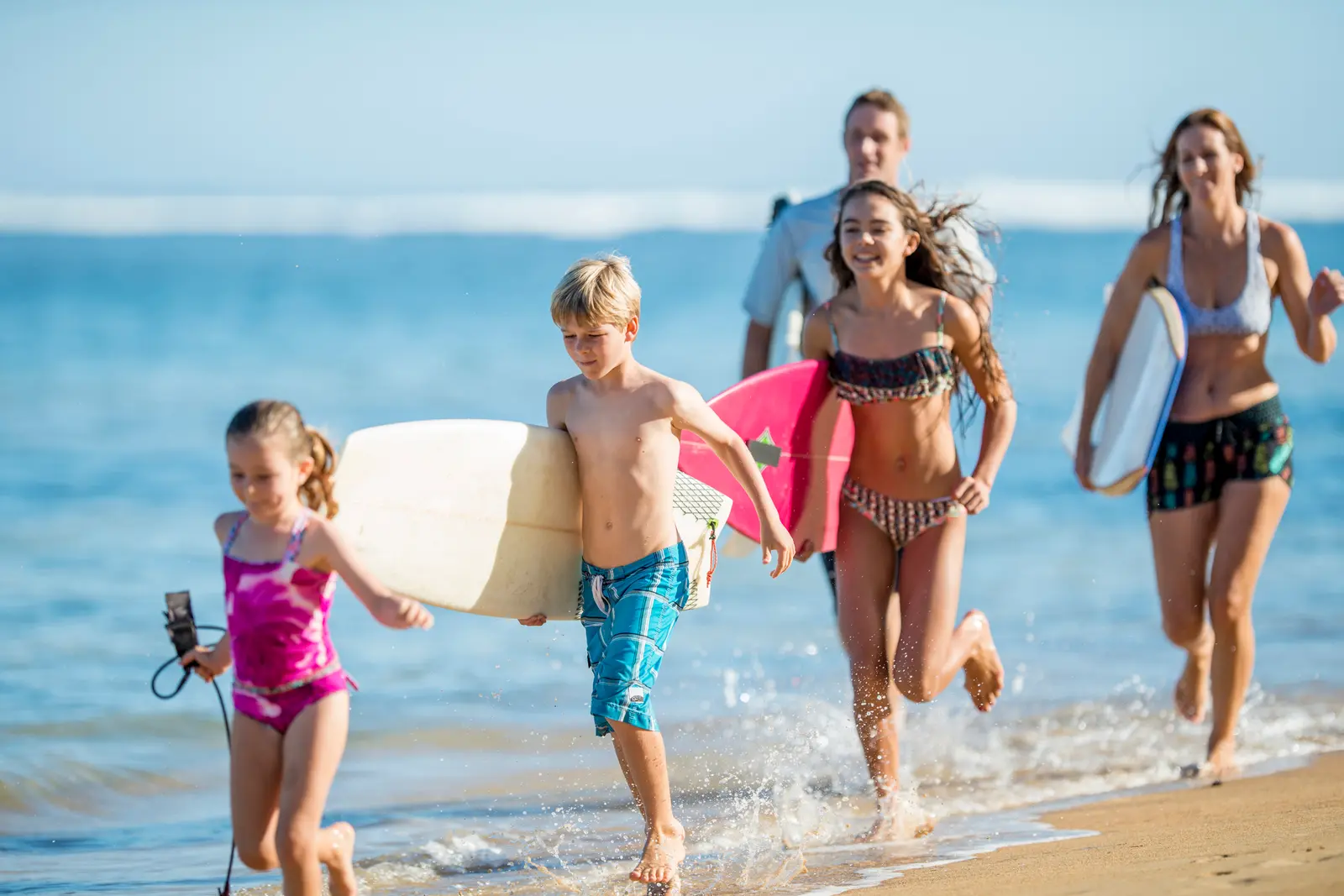 Beach days with the family. Four people in swimwear walk along the wet sand carrying surfboards, with the ocean and a blue sky behind them.
