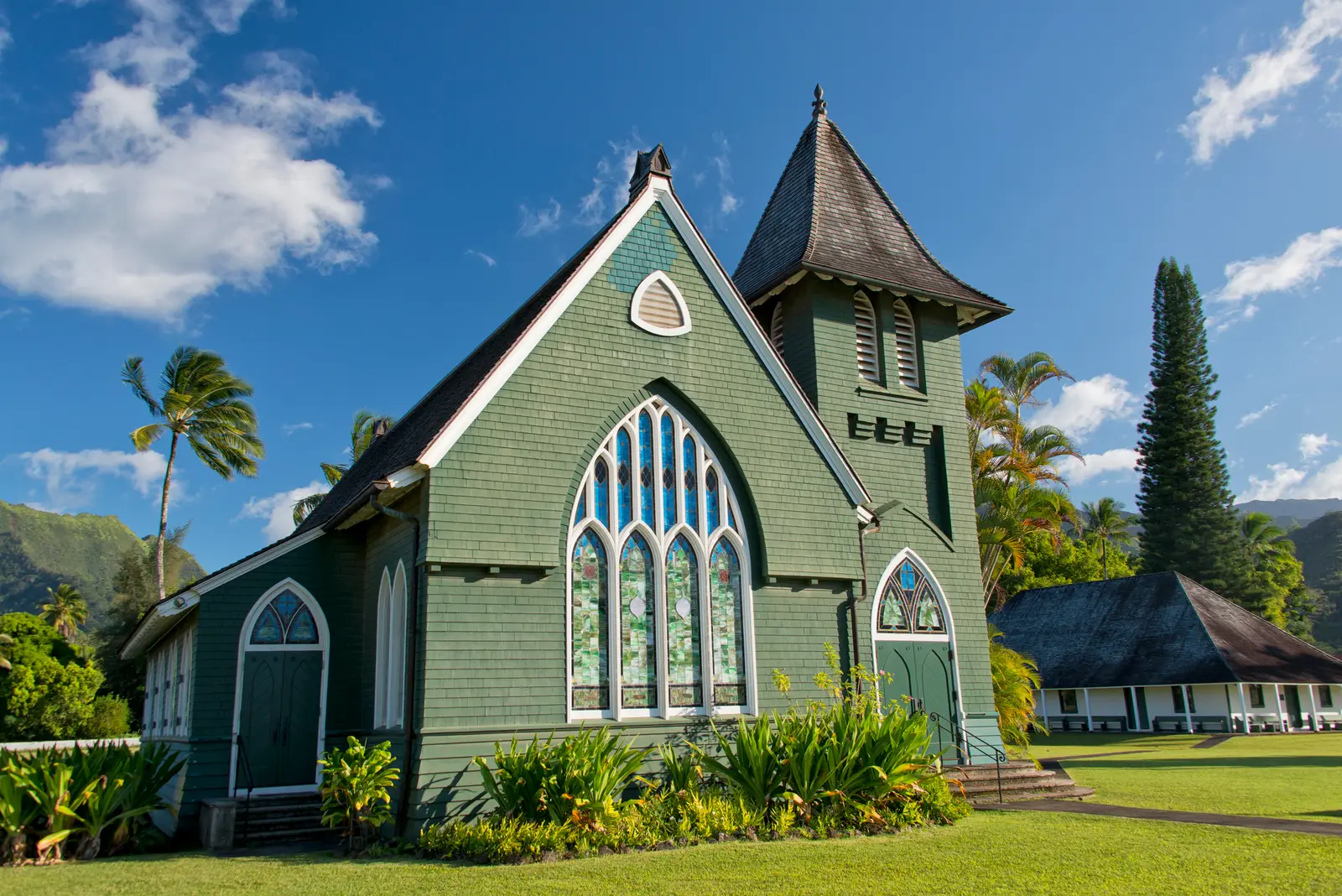 Historic church in Hanalei. A green wooden church with white trim and a steeple, seen through palms and a white picket fence.