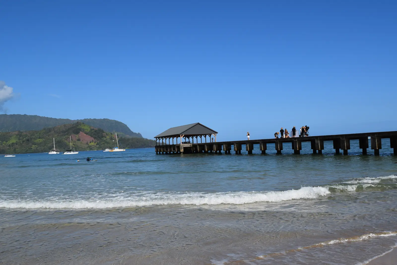 Hanalei Pier on Hanalei Bay. A long pier with a covered shelter at the end stretches into calm water, with mountains along the far shore.