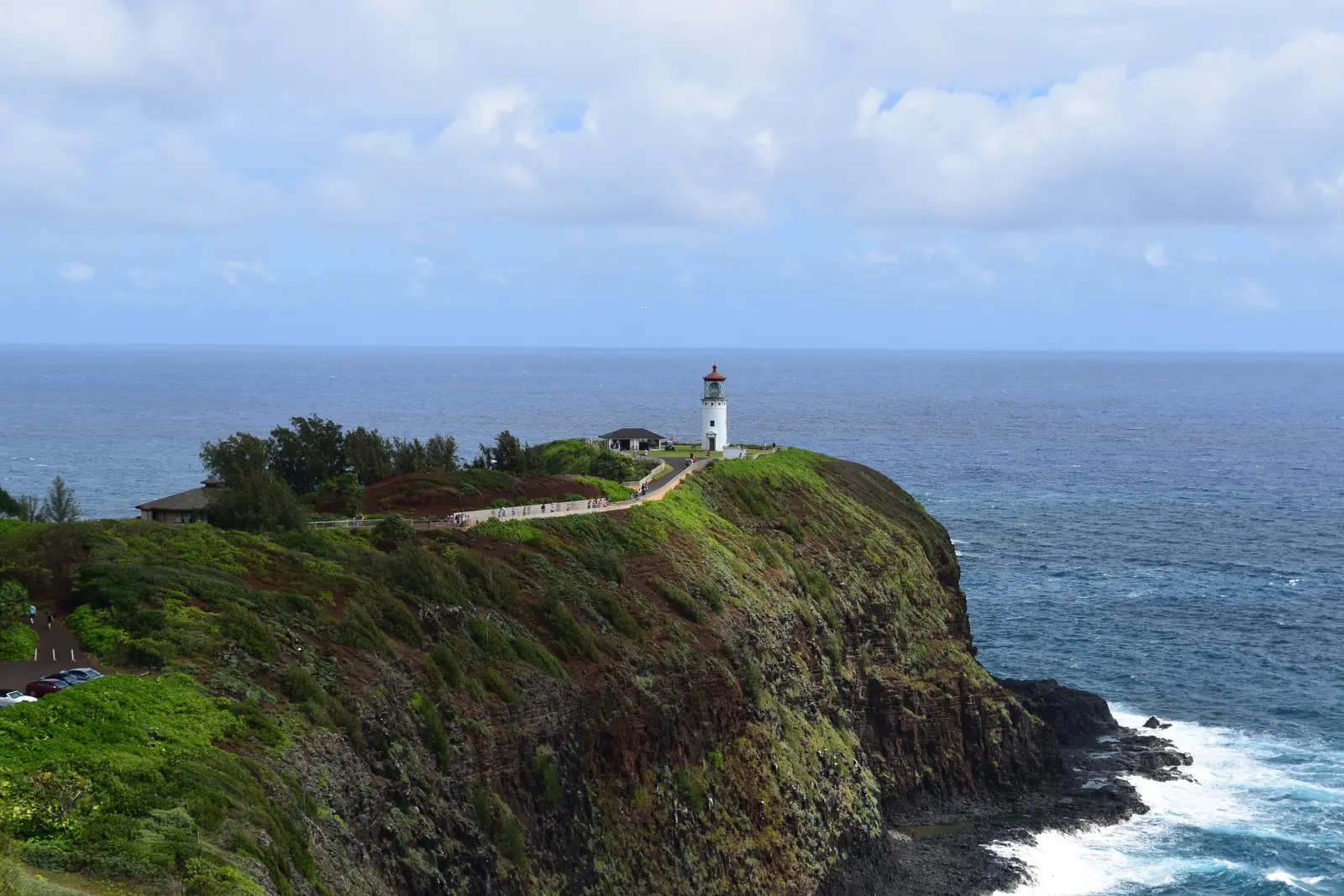 Kīlauea Lighthouse. A white lighthouse stands on a grassy cliff above the ocean, with green hills behind it.