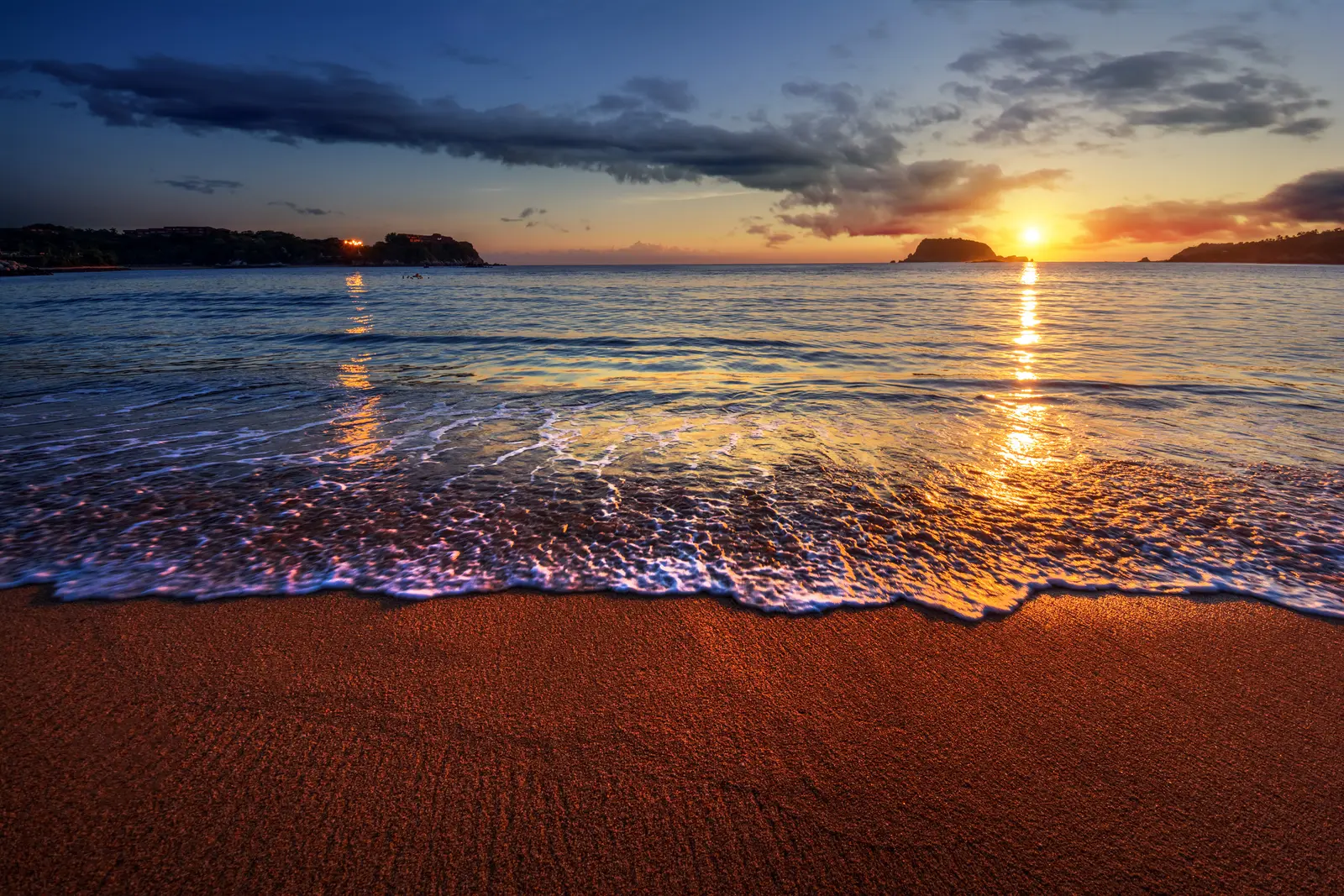 Sunset on the beach. People stand and sit on the sand while the sky turns orange over the water.