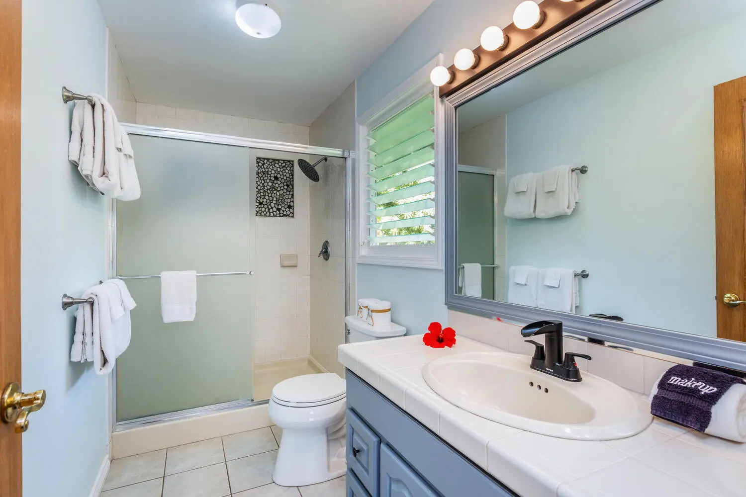 Bahama bathroom. A glass-enclosed shower, a toilet, and a blue-gray vanity with an oval sink, a lit mirror, white towels, and a shuttered window onto foliage.
