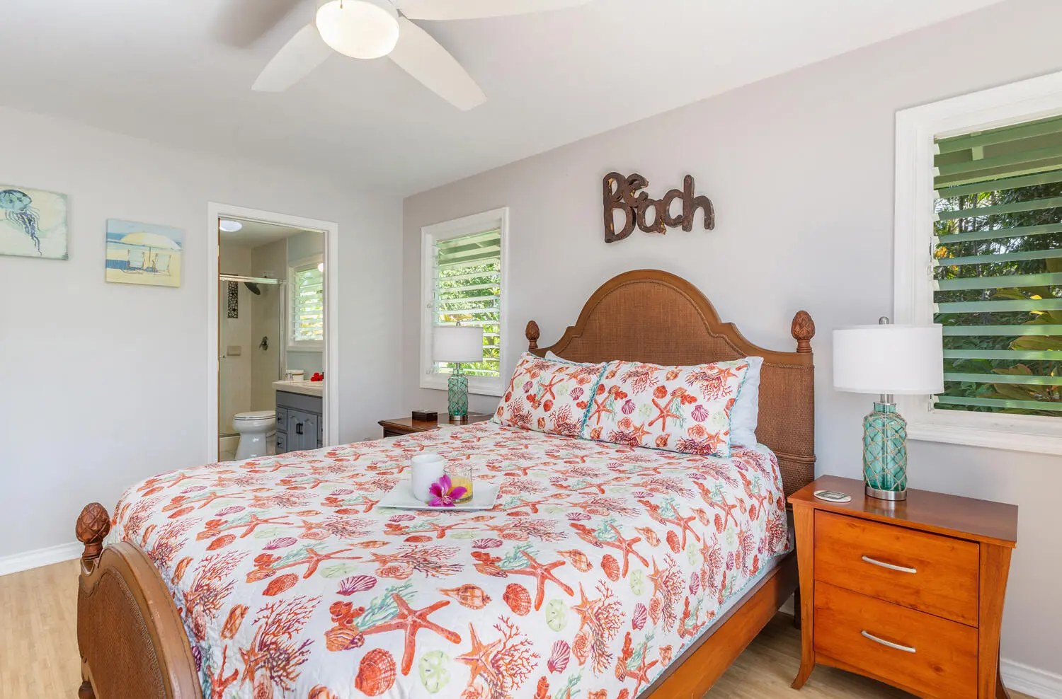 Bahama bedroom. The cane-headboard bed and starfish quilt, turquoise lamps, shuttered windows onto foliage, and an open doorway into the bathroom.