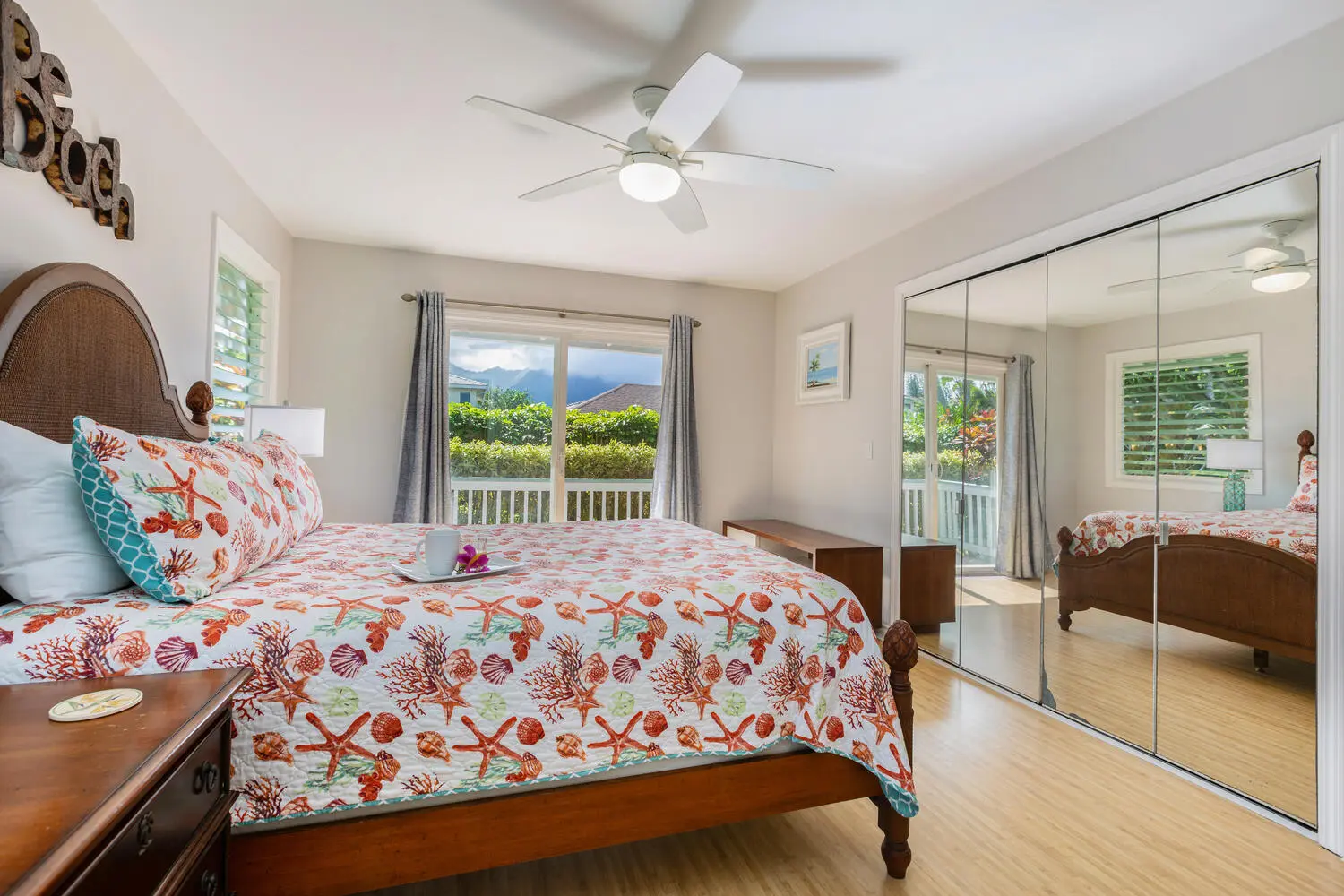 Bahama bedroom. A wood bed with a cane headboard and a white quilt printed with starfish, shells, and coral, mirrored closet doors, and a sliding door toward a balcony and mountains.