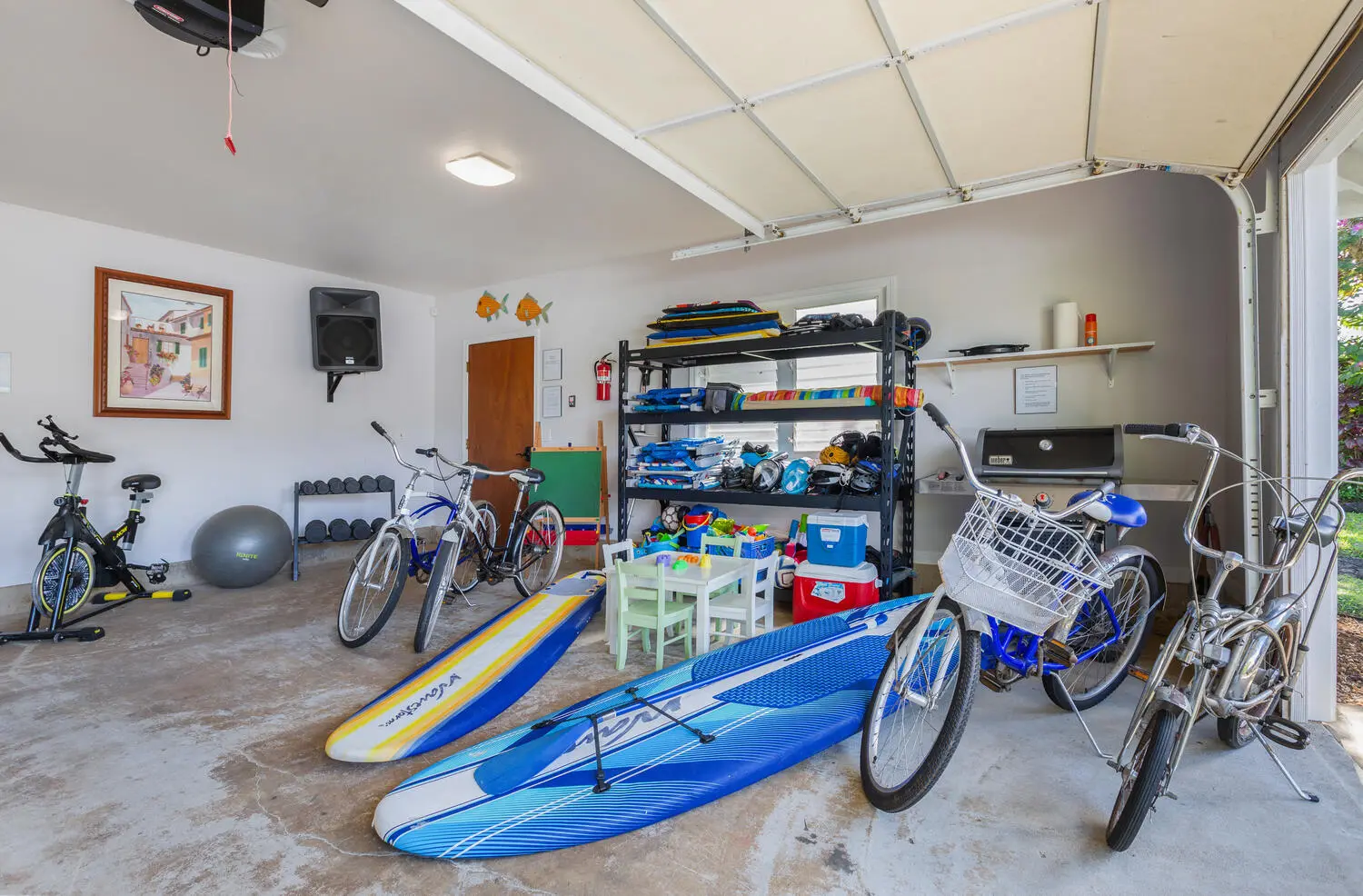 Beach toys, bikes, and exercise area. An open garage with stand-up paddleboards, bicycles, a stationary bike, an exercise ball, a dumbbell rack, and shelves of pool and beach gear.
