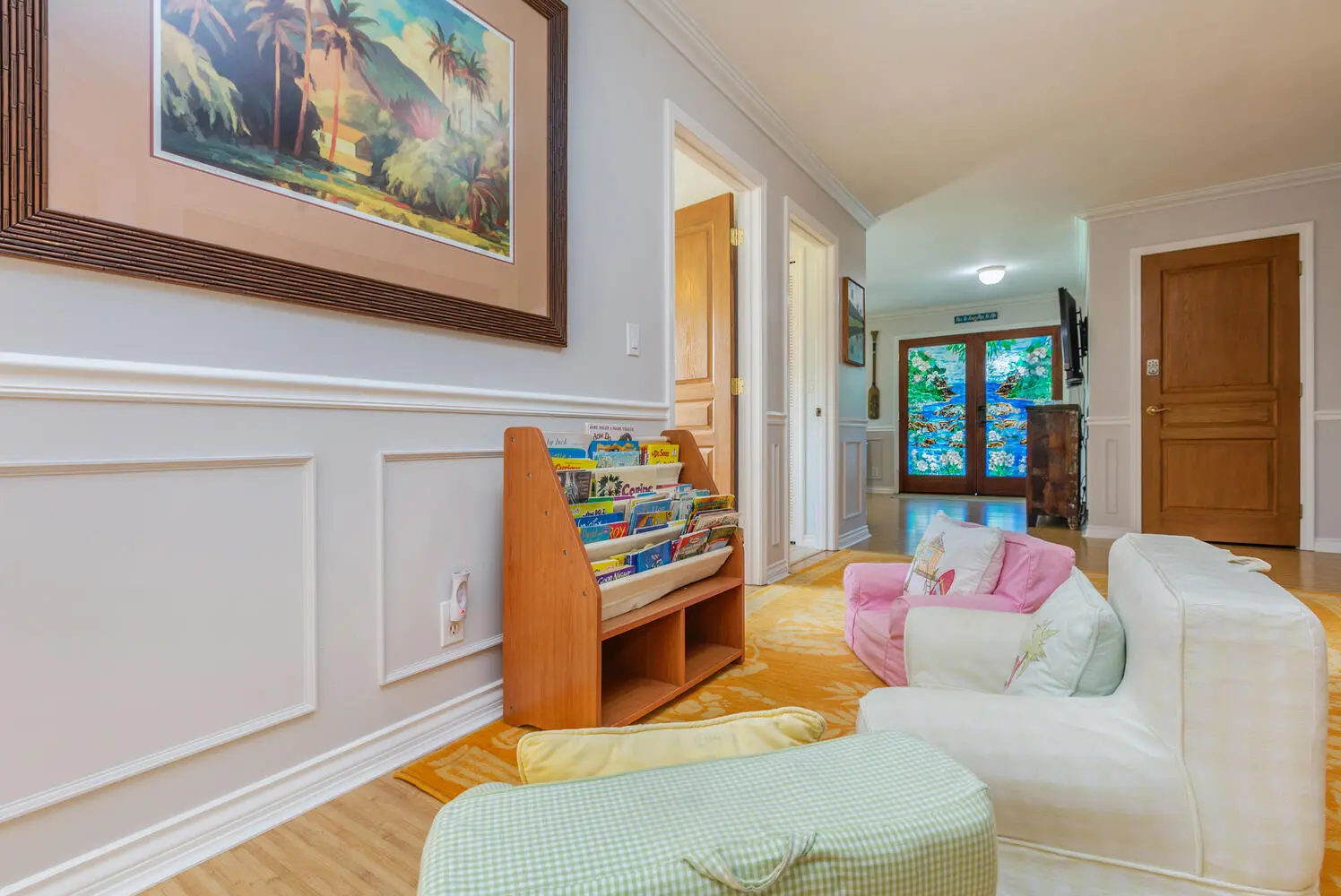 Children's reading area. A white seat, a pink child's chair, floor cushions, and a rack of children's books on a yellow rug, beside a tropical painting and stained-glass doors.