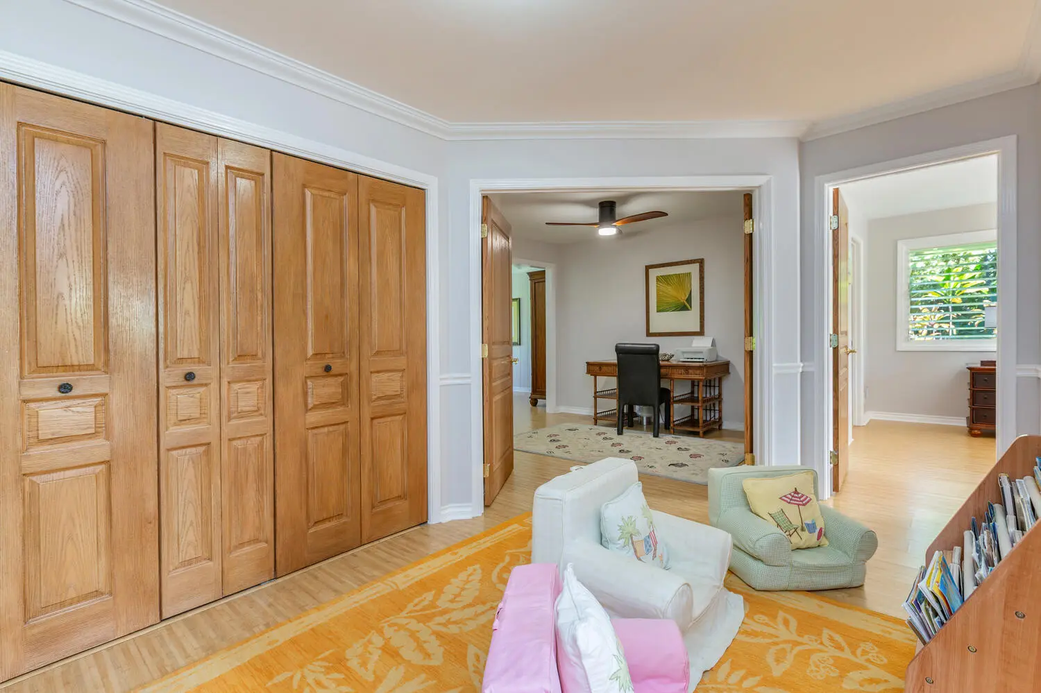 Children's reading area. A white chair, a small green child's chair, and books on a low shelf, with light oak closet doors and a doorway into a room with a desk.
