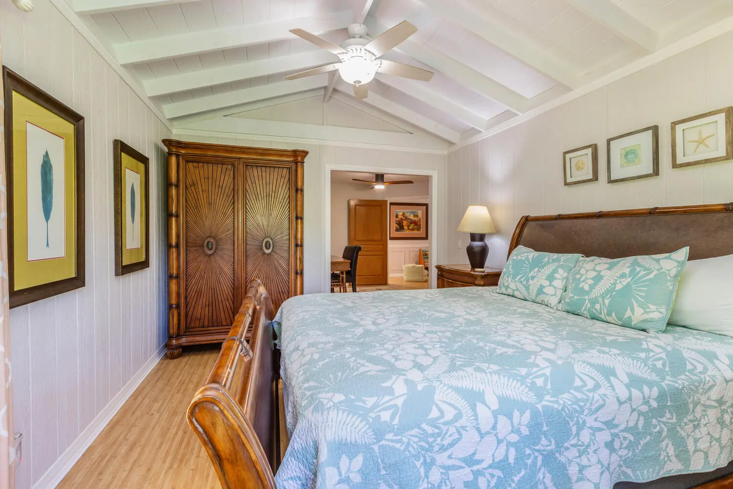 Cottage bedroom. A bed with a bamboo-style wood headboard and a light blue and white leaf quilt, a carved armoire, light wood floors, and a vaulted ceiling with a fan.