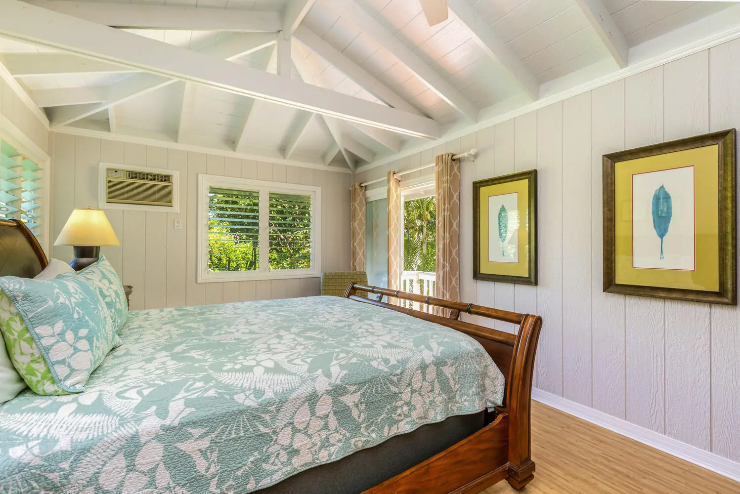 Cottage bedroom. A dark wood sleigh bed with a teal-and-white leaf quilt, a vaulted beamed ceiling, shuttered windows onto foliage, and a glass door toward palms.