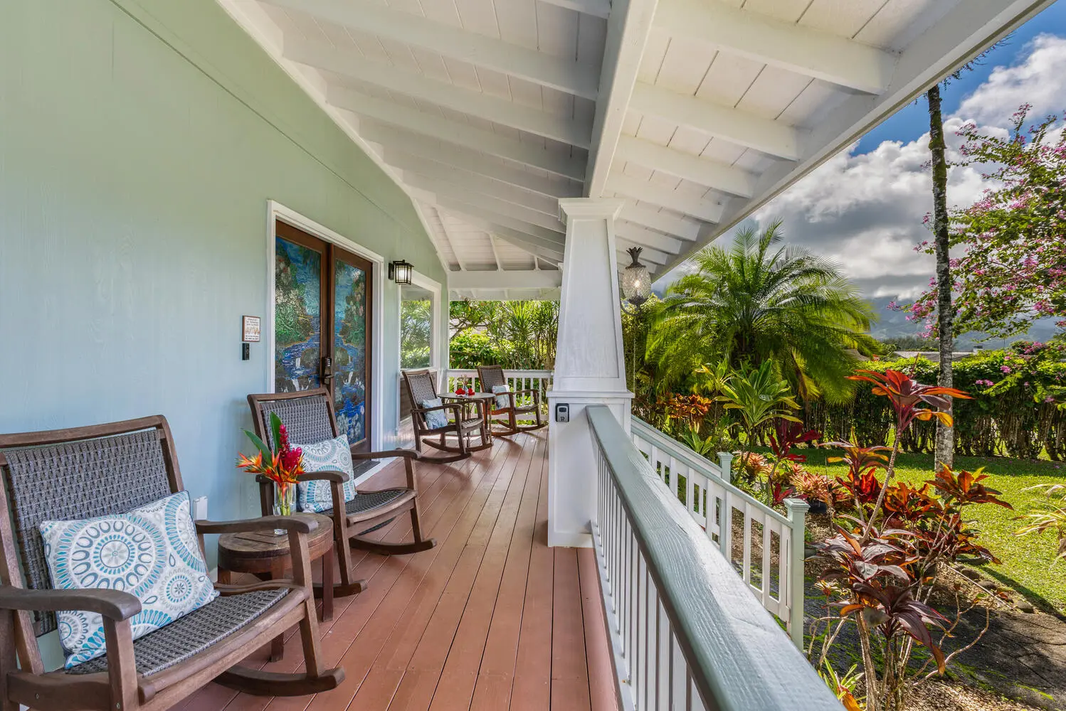 Front lanai with rocking chairs. A covered lanai with a wood deck, a white beamed ceiling, rocking chairs with blue-and-white cushions, and a view of the lawn, palms, and mountains.