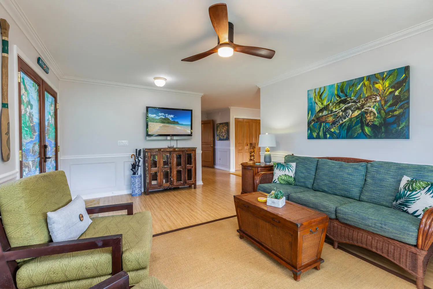 Entry living area. A teal sofa and a green armchair around a wooden chest, a wall-mounted television, a sea-turtle painting, and light wood floors.