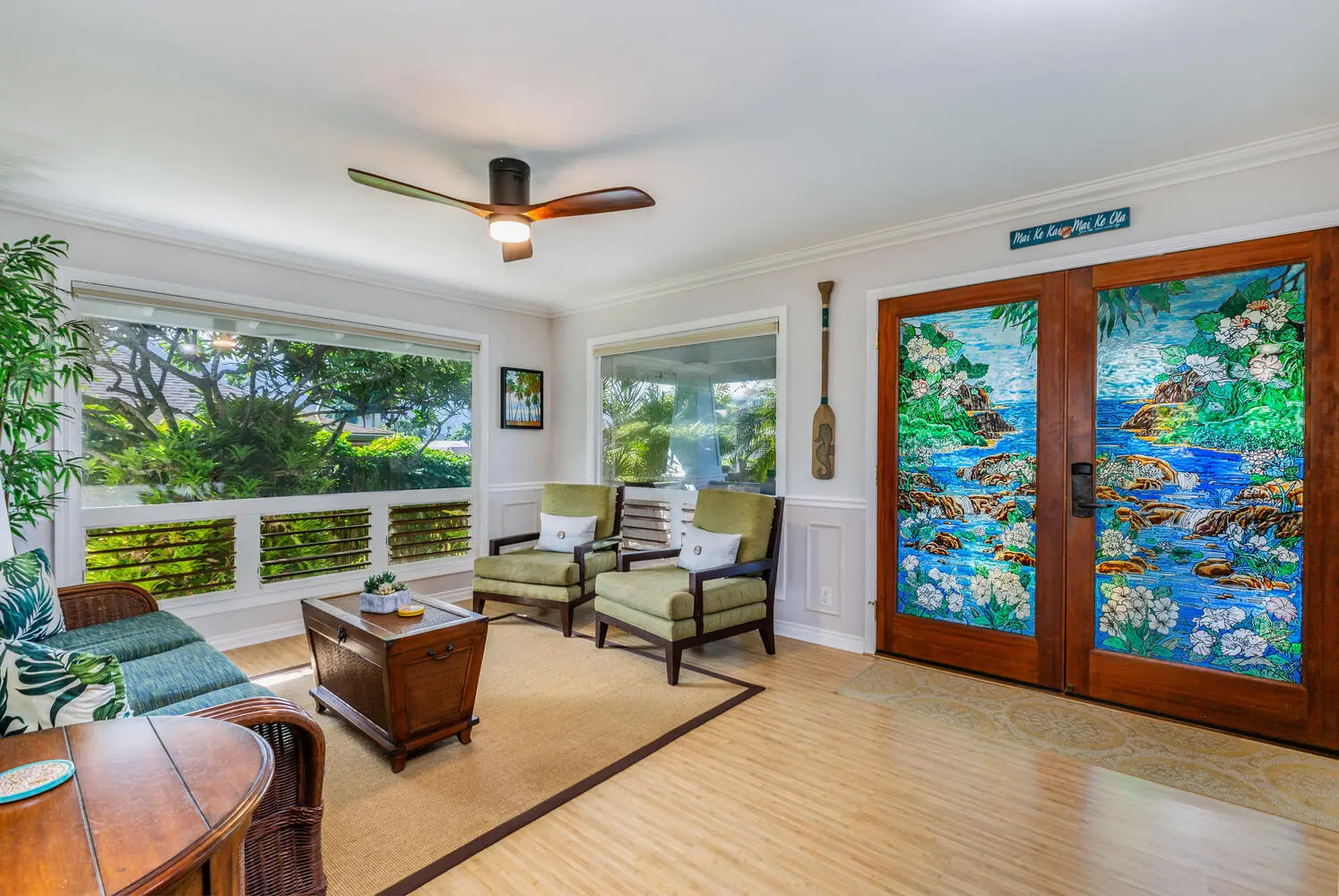 Entry living area. Green armchairs and a blue-green sofa around a wooden trunk, light wood floors, a ceiling fan, and windows onto the garden, with stained-glass doors.