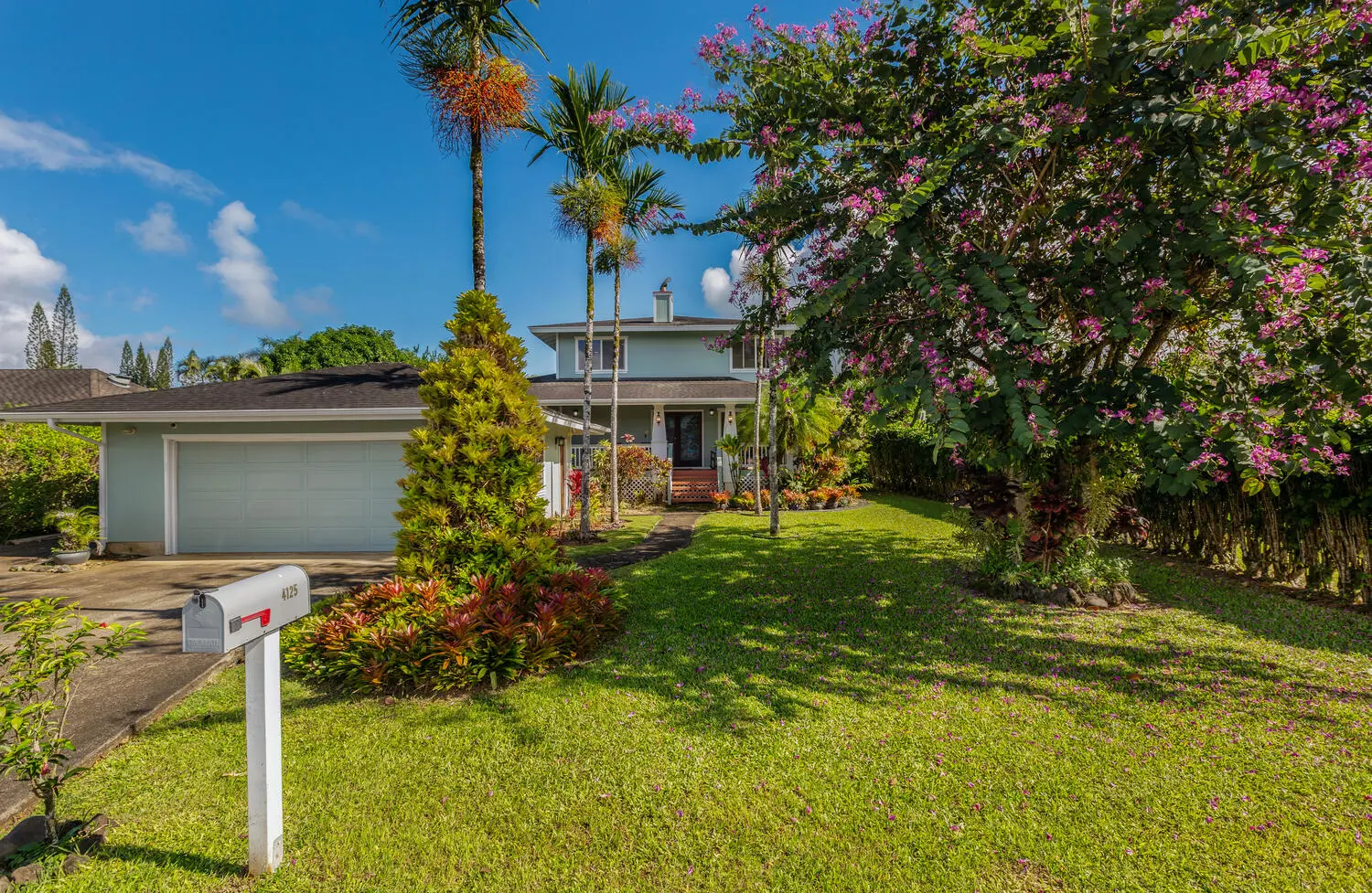 Front of Lani Hanalei Home. The house across the lawn, with a garage door and driveway, palms, flowering trees, and the porch in the distance.
