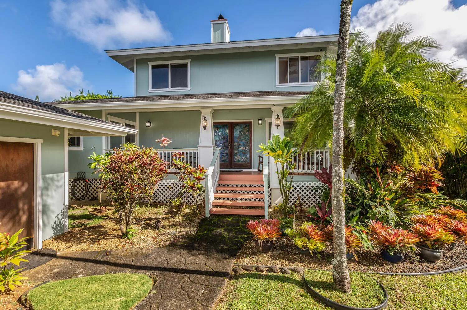 Front of Lani Hanalei Home. A two-story house with light aqua siding, a covered porch with white columns and railings, wood stairs, stained-glass doors, and tropical plants.