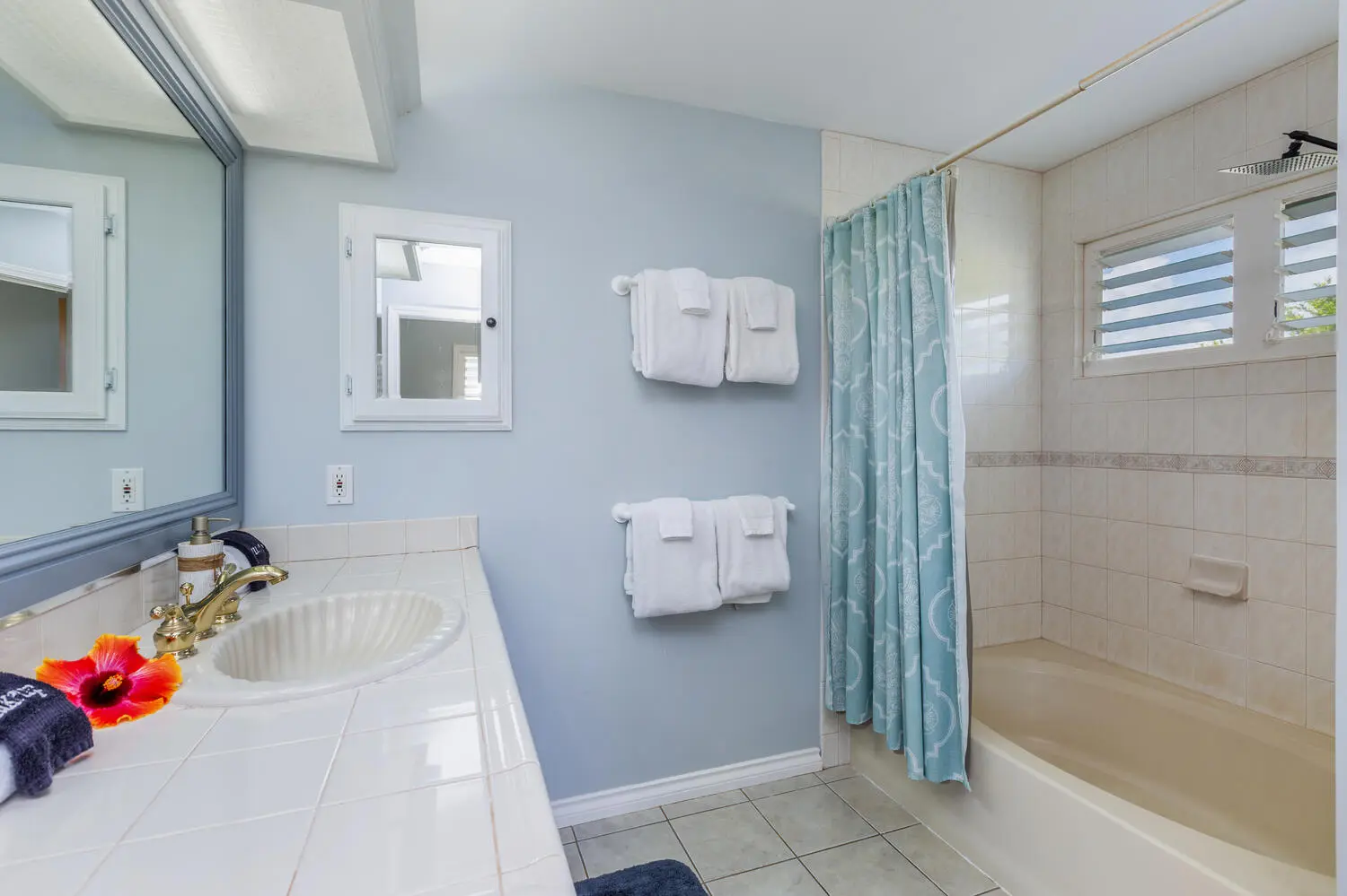 Lani Hanalei Suite bathroom. A built-in tub with a light blue shower curtain, a shell-shaped sink on a tiled counter, towel racks, and a louvered window above the tub.