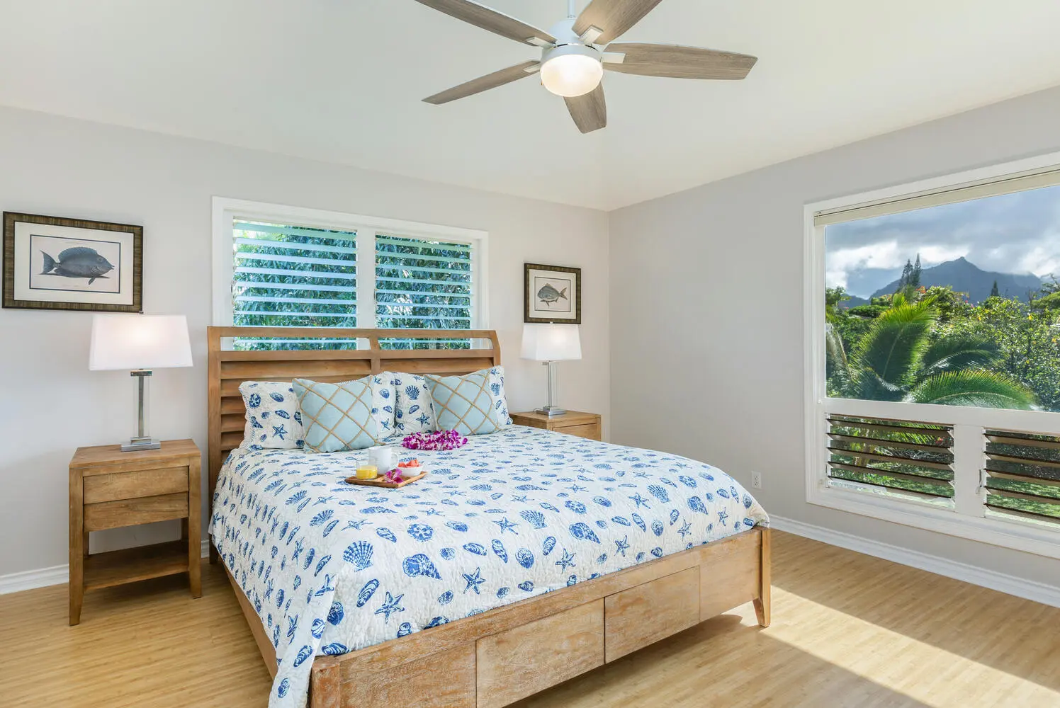 Lani Hanalei Suite. A wood bed with a white quilt printed with blue shells, starfish, and coral, fish prints on the walls, louvered windows onto foliage, and a wide window toward mountains and palms.