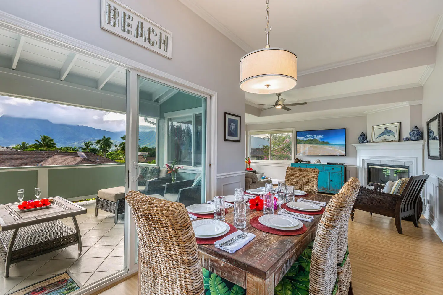 Main dining area and lanai. A rustic wood table and woven chairs open through a sliding glass door to a covered lanai with wicker seating, mountains, and palms. A fireplace and a television are on the far wall.