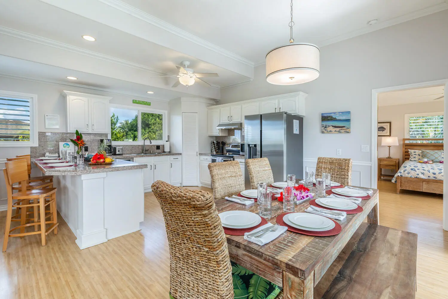 Main dining area - upstairs. A rustic wood dining table set with white plates and red placemats, woven chairs, and a drum pendant, with the kitchen island nearby.