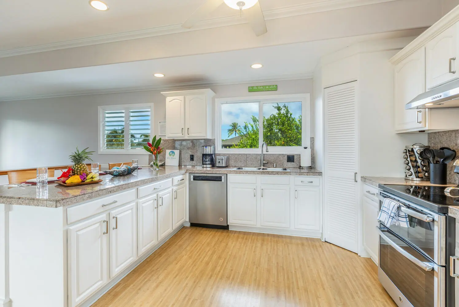 Main kitchen. White cabinets and granite counters, a double sink under a window looking at trees and a palm, and a stainless gas range beside a louvered door.