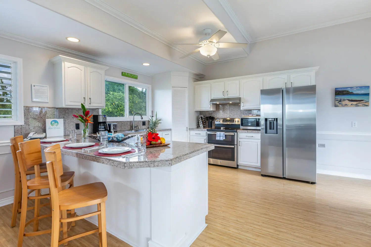 Main kitchen - upstairs. White cabinets, a granite island set with plates and wooden bar stools, and a stainless refrigerator, range, and microwave, with trees outside the window.