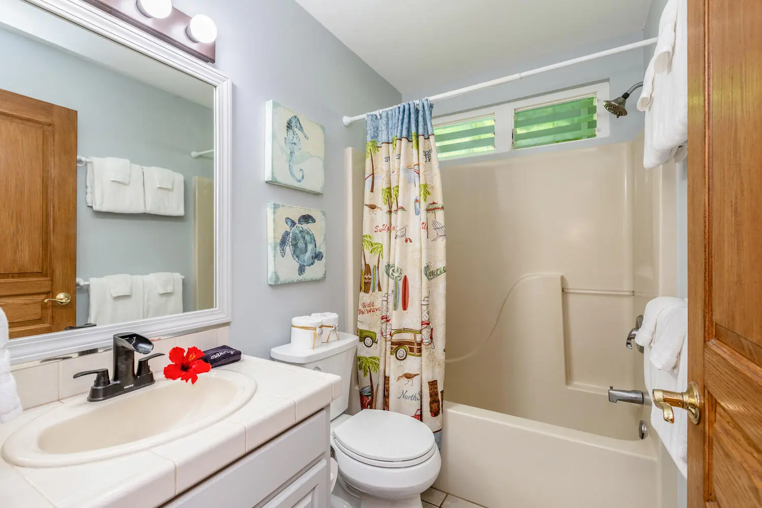 ʻOhana bathroom. A white vanity and sink beside a toilet and a bathtub with a tropical-print curtain, seahorse and sea-turtle pictures, and narrow windows with green blinds.