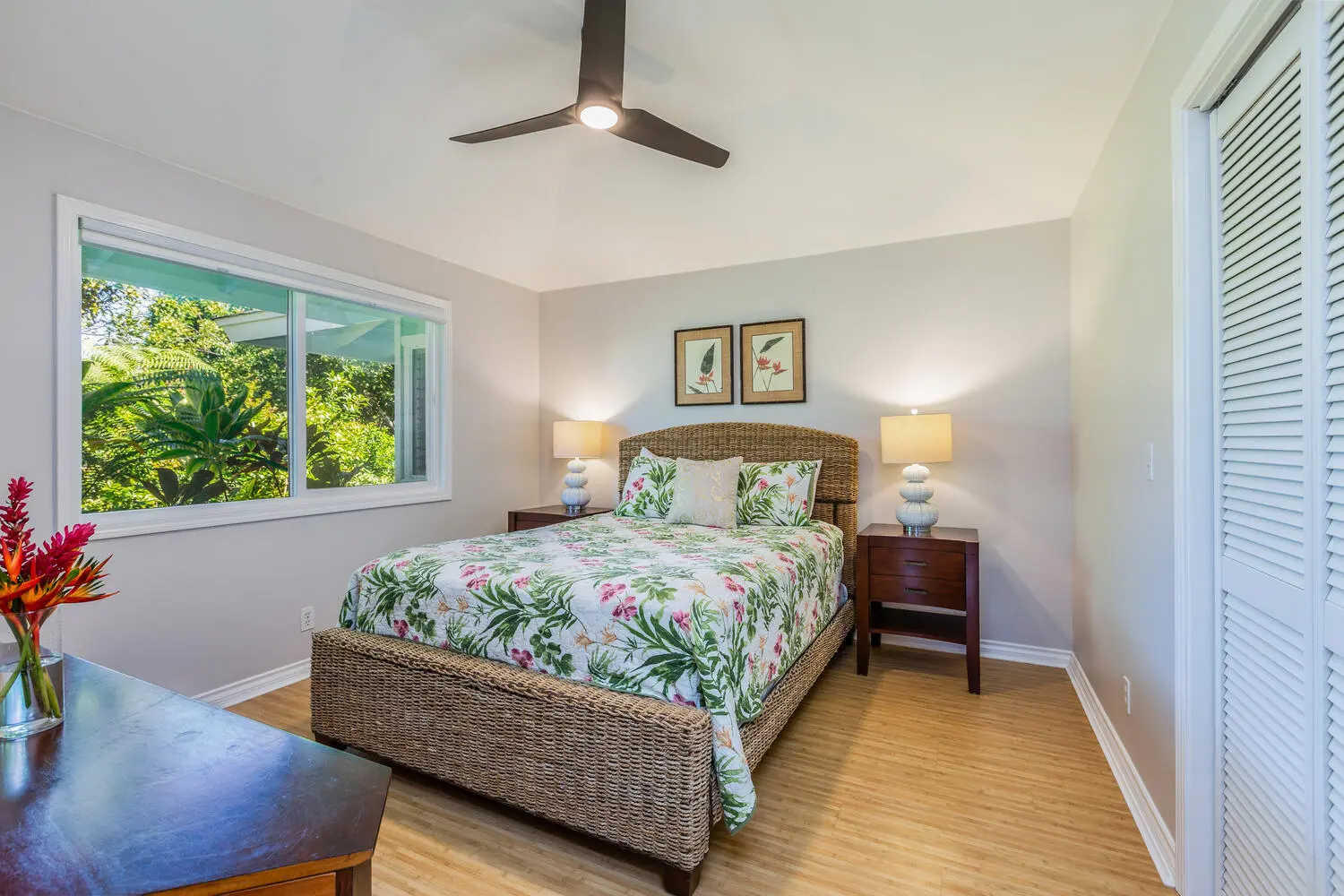 ʻOhana bedroom. A bed with a woven wicker headboard and a white quilt with green and pink flowers, dark wood nightstands, louvered closet doors, and a window onto the garden.