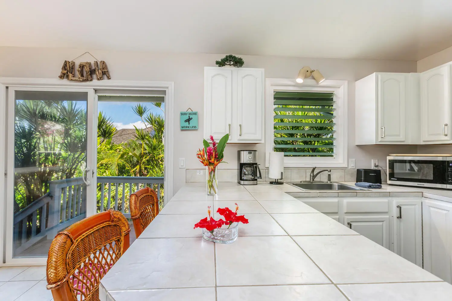 ʻOhana kitchen. White cabinets, a tiled island with two wicker chairs, a sink and microwave, and a sliding door onto a deck with palms and garden.