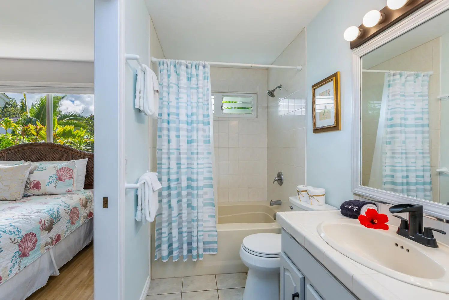 Pineapple bathroom. A bathtub with a blue-and-white striped curtain, a toilet, and a white vanity, with a bedroom and its seashell quilt visible through the doorway.