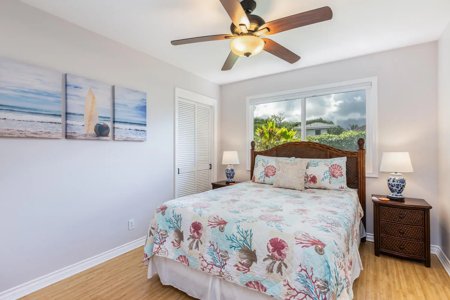 Pineapple bedroom. A wood bed with a cane headboard and a shell-and-coral quilt, dark wood nightstands, white louvered closet doors, beach paintings, and a window onto tropical plants.