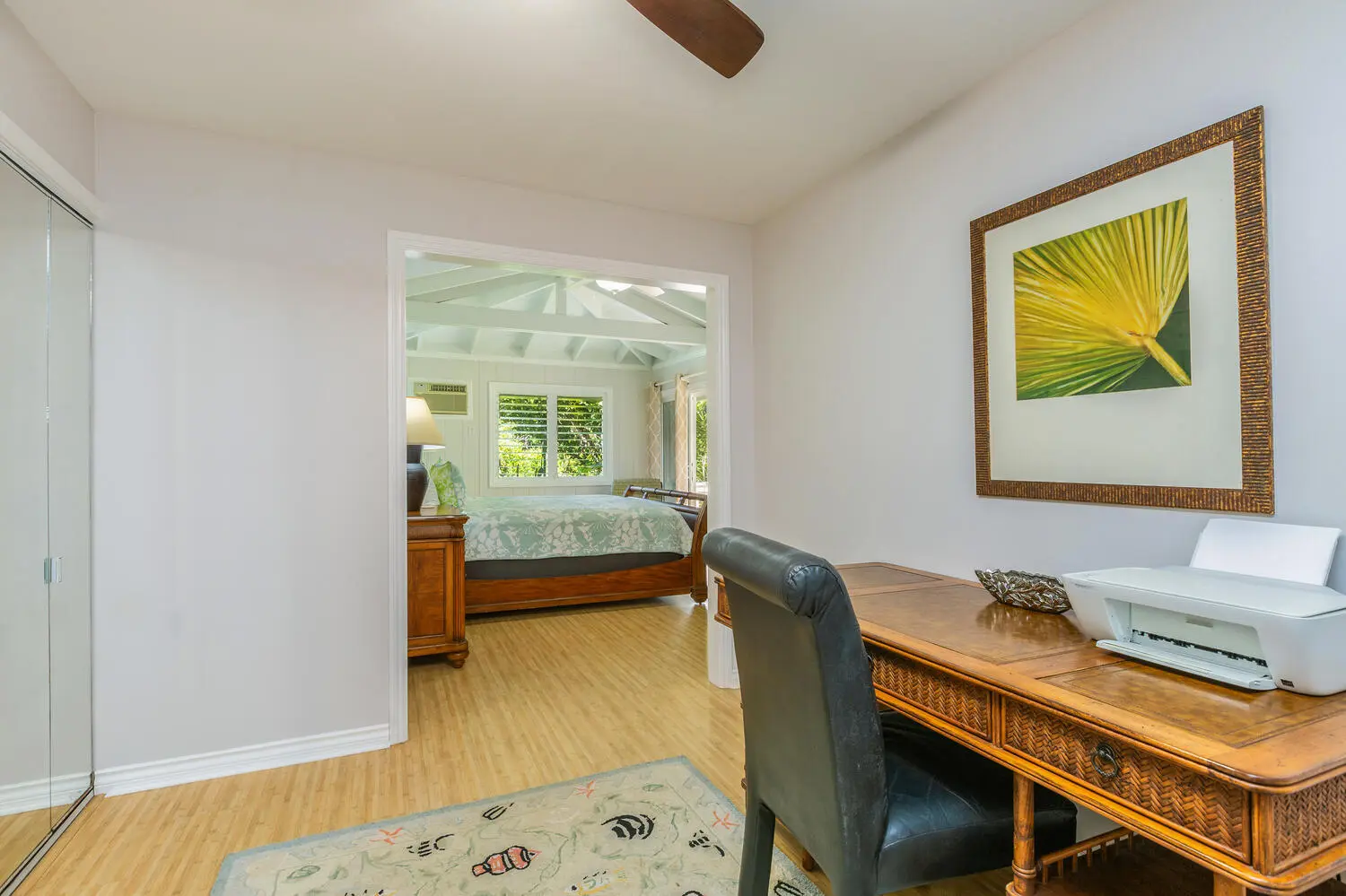 Study area. A wooden desk, black chair, and printer facing a palm-frond print, with mirrored closet doors, and a bedroom with a leaf-patterned quilt visible through the doorway.