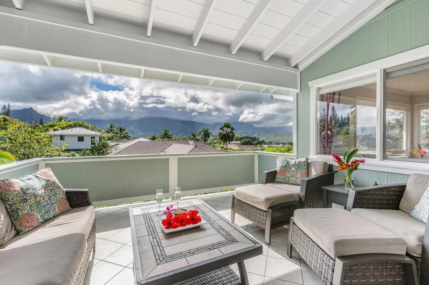 Upstairs lanai. Wicker sofas and chairs with beige cushions around a tile-top table, a tiled floor, pale green walls, and an open view of green mountains and palms.