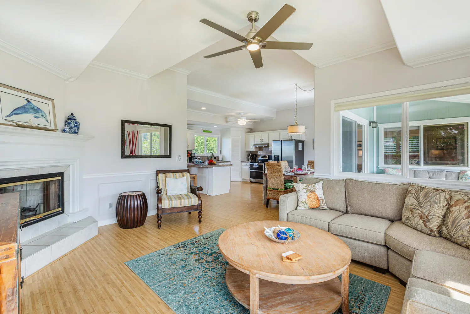 Upstairs living area. A beige sofa, round coffee table, striped armchair, and white fireplace, opening to white kitchen cabinets, an island, and a dining table, with windows toward covered outdoor seating.