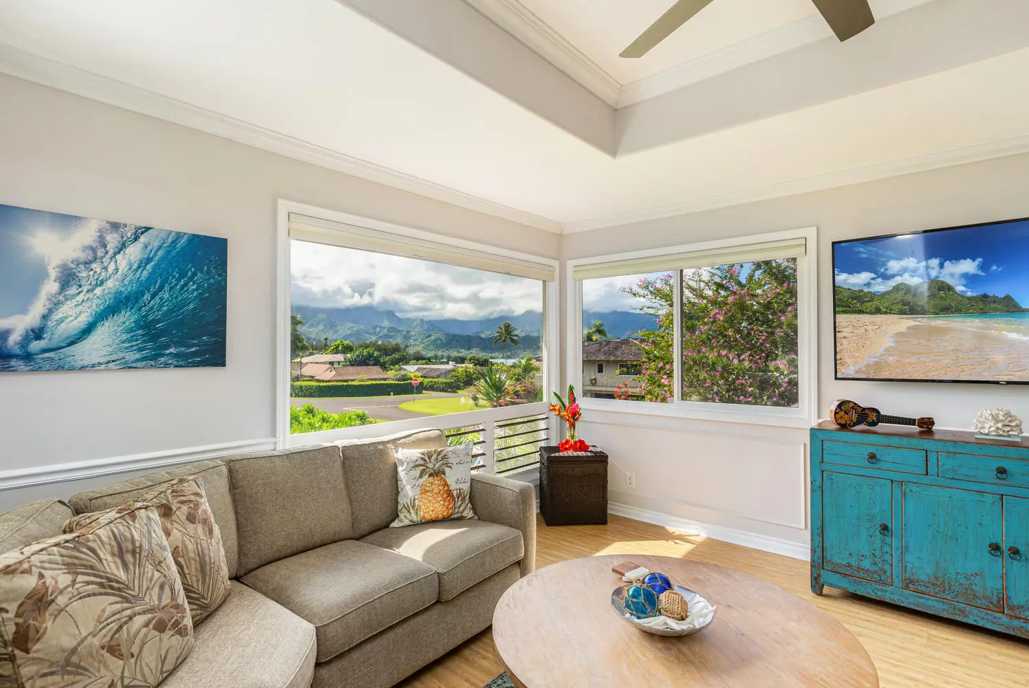 Upstairs living area. A beige sofa with leaf-patterned cushions beside a round wood table, a turquoise cabinet and television, and windows toward mountains, palms, and a white railing.