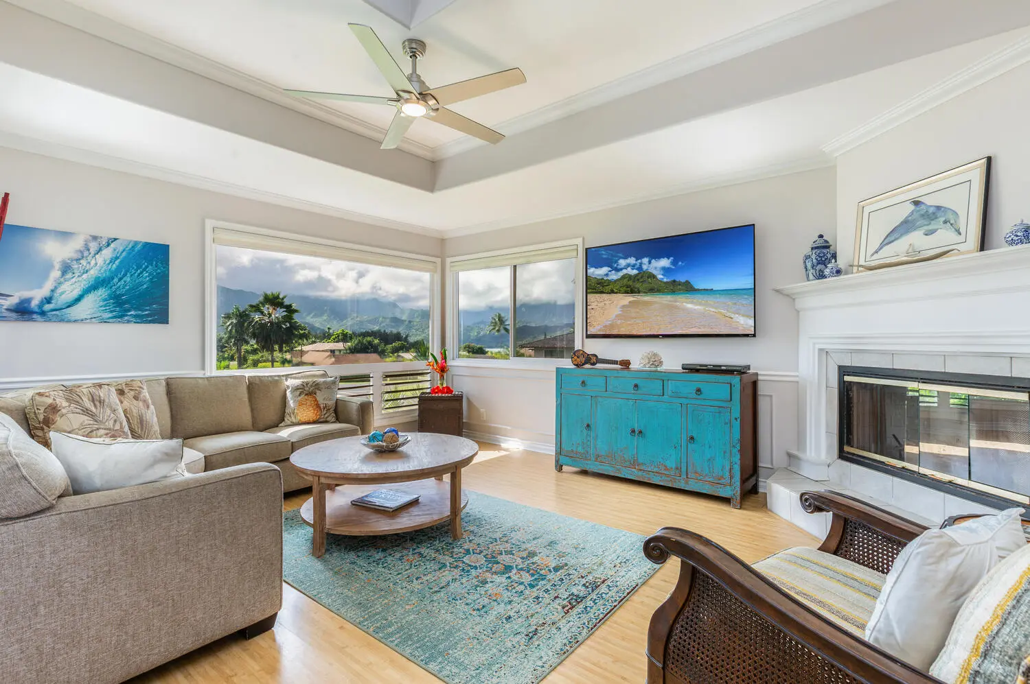Upstairs living area. Beige sofas around a round wood coffee table, a turquoise cabinet under a television, a white fireplace, and windows looking out to green mountains and palms.